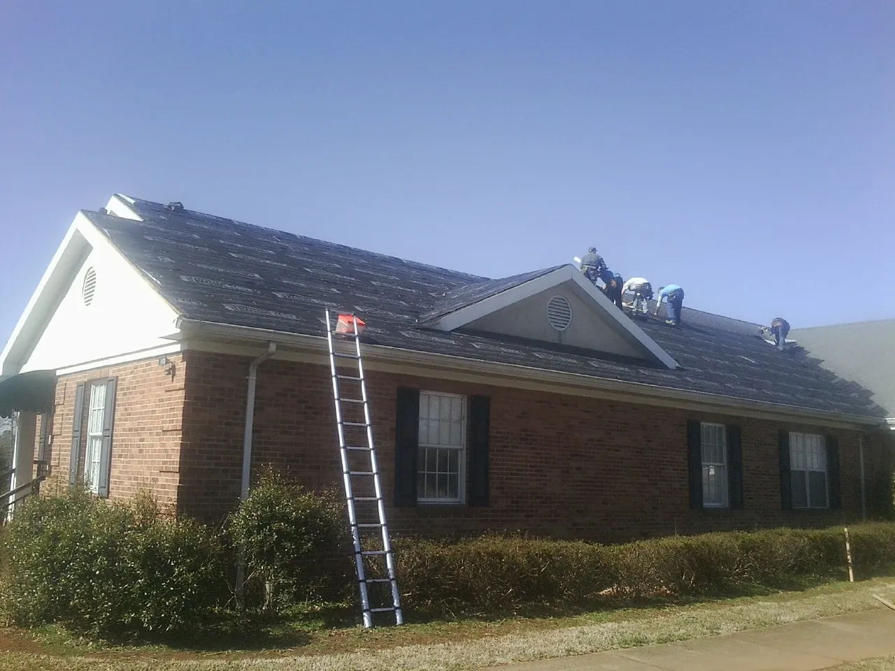 Crew tearing shingles off a brick ranch roof with a ladder set — Emergency Roof Repair work by our crew in Ramseur