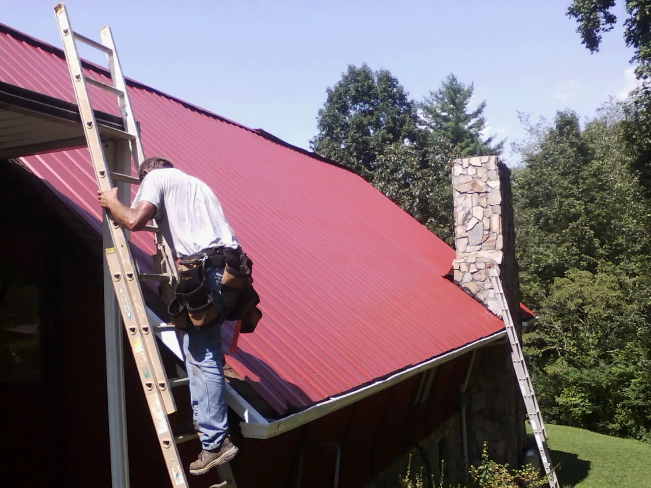 Roofer on a ladder beside a red metal roof and stone chimney — Soffit & Fascia Repair by Roofing Brothers in Ramseur